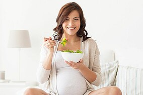 Schwangere sitzt auf einem Sofa und isst einen Salat